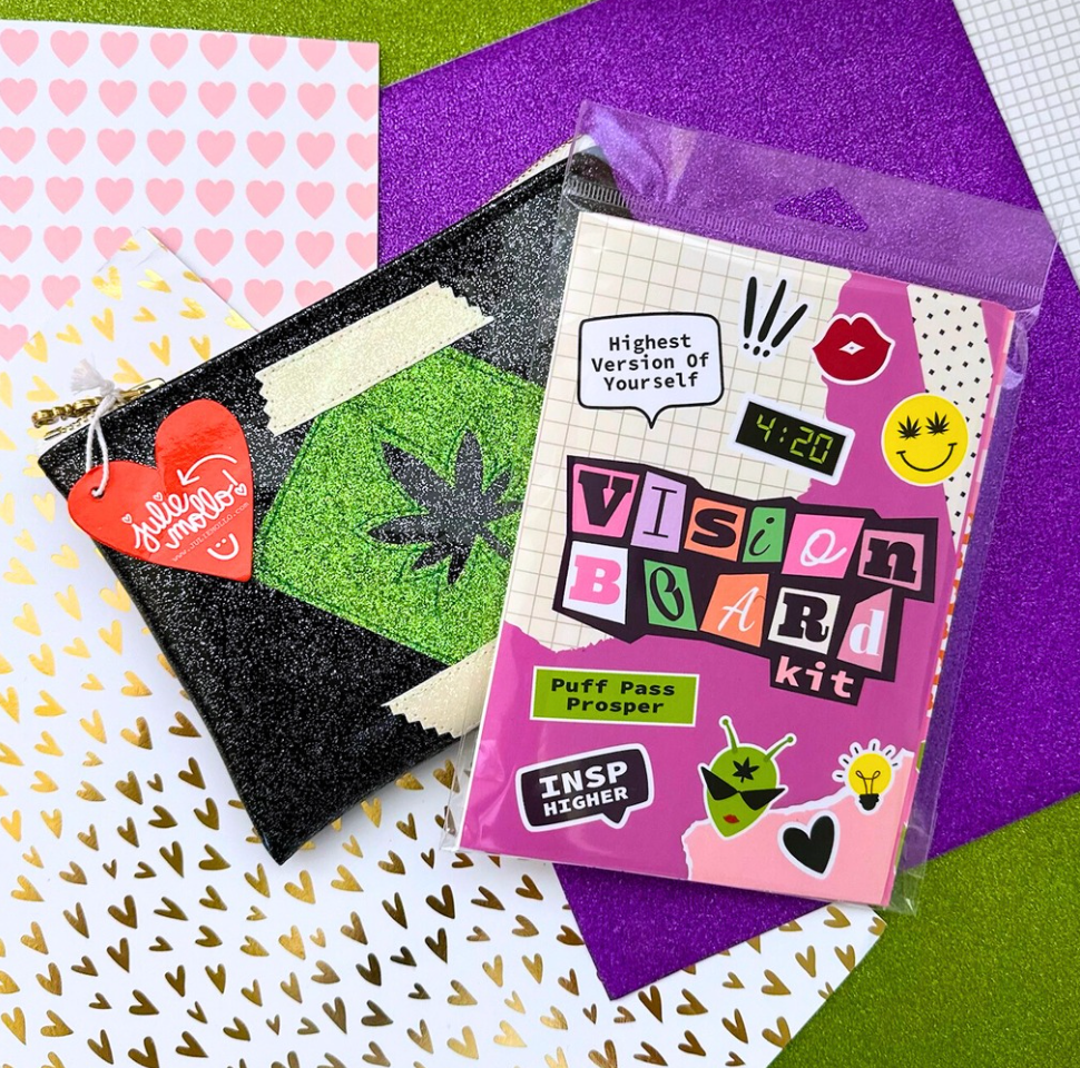 Vision Board kit with colorful stickers on a patterned surface with A black glittery pouch with a green pot leaf cut out of the center. The pouch has a zipper closure and a red heart-shaped tag that reads "Julie Mollo" attached to the zipper pull. It rests on a wooden surface.