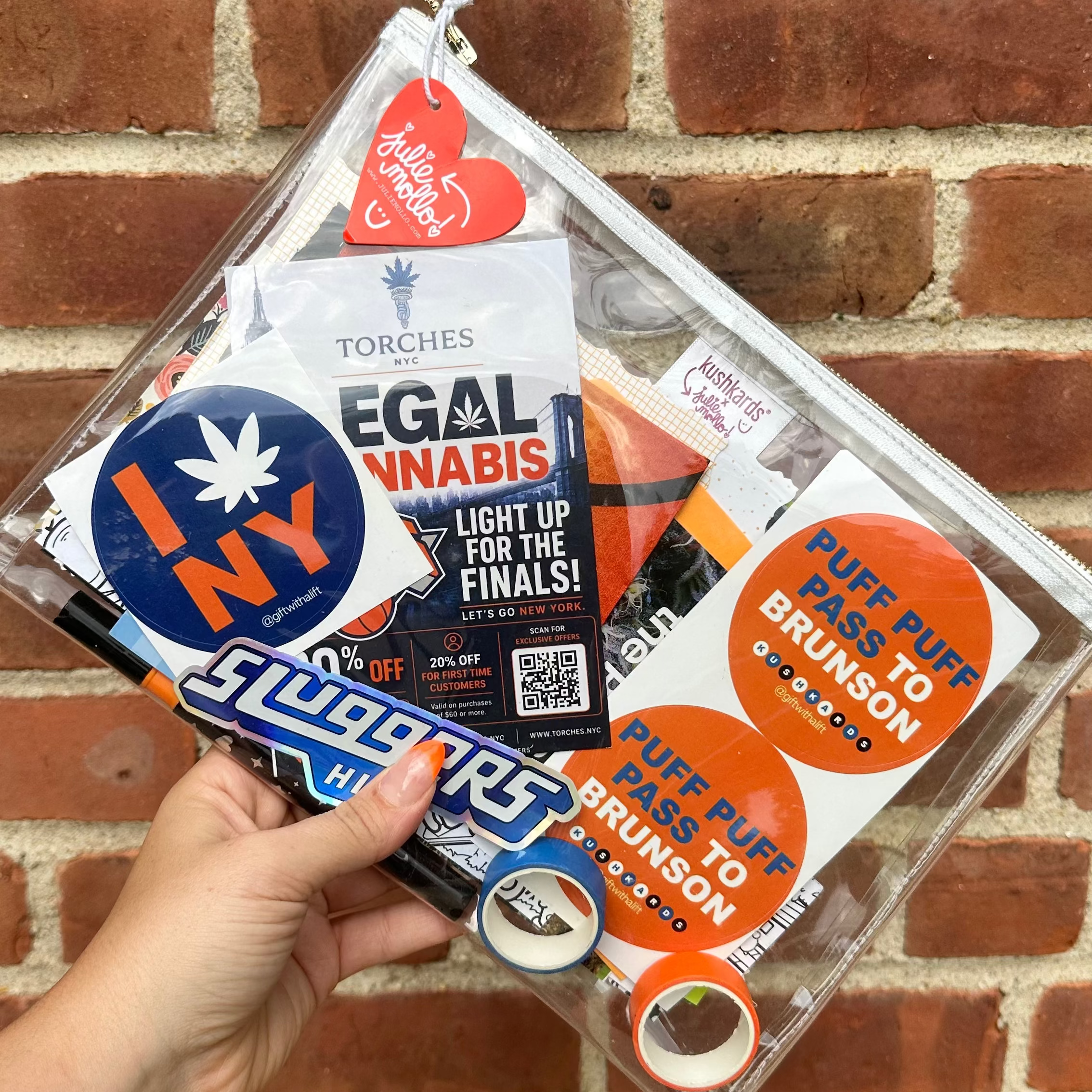 Hand holding a clear clutch bag with various stickers against a brick wall.