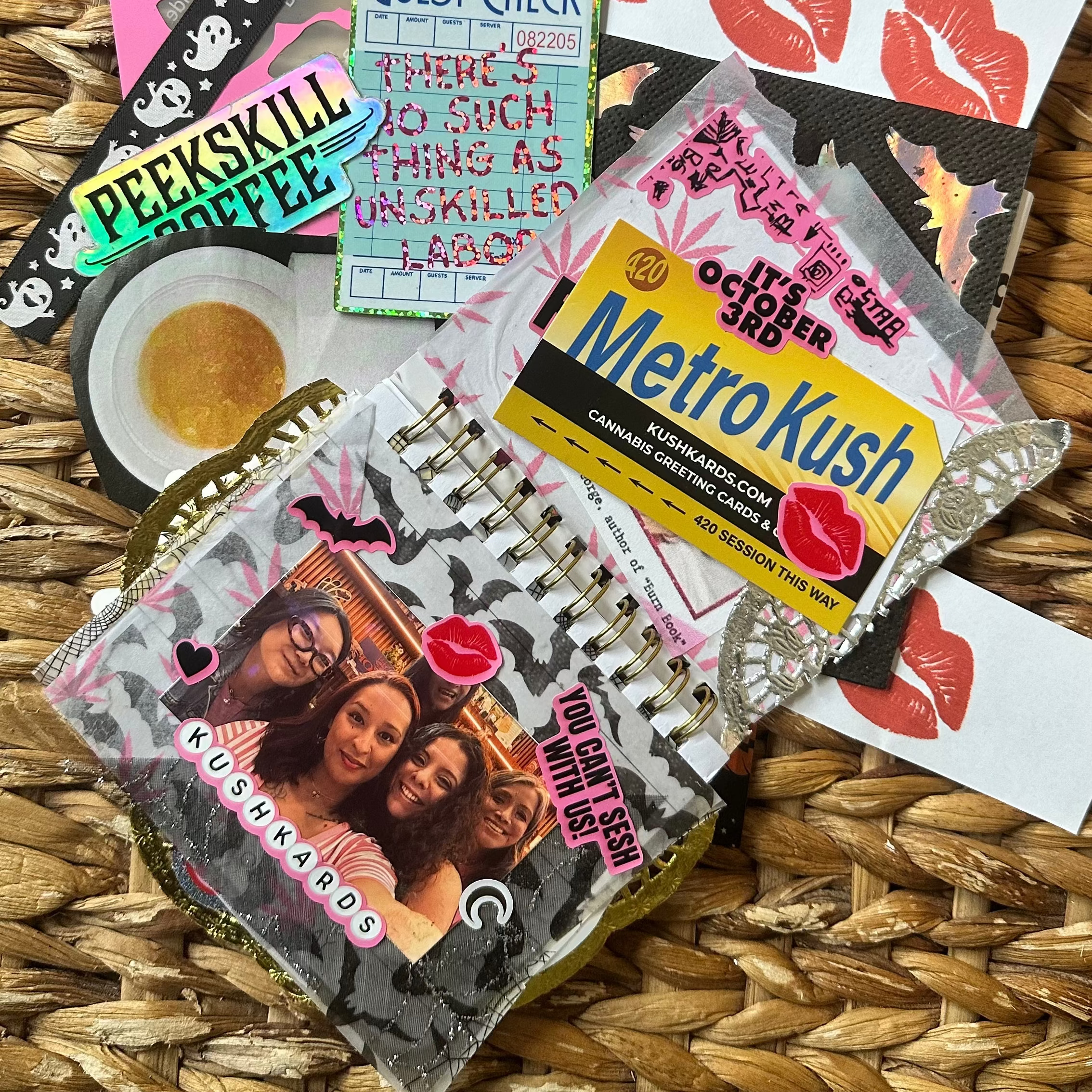 A curated assortment of themed stationery and accessories laid flat on a woven straw mat, including a pink lip-print notebook, a black-and-white skull-patterned ribbon, a &