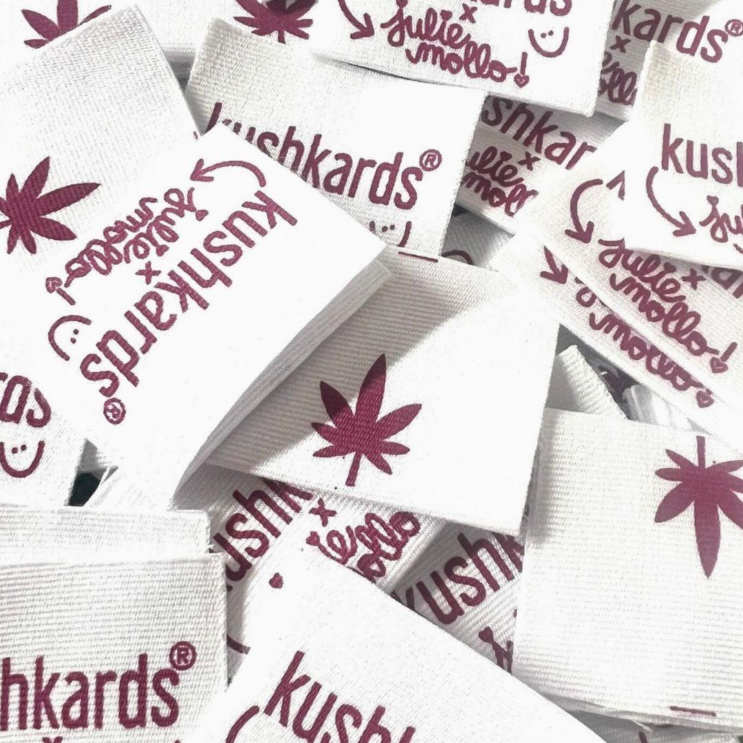 Stack of white fabric labels featuring the KushKards x Julie Mollo collaboration, printed with a maroon cannabis leaf and playful typography, designed for the collaboration collection
