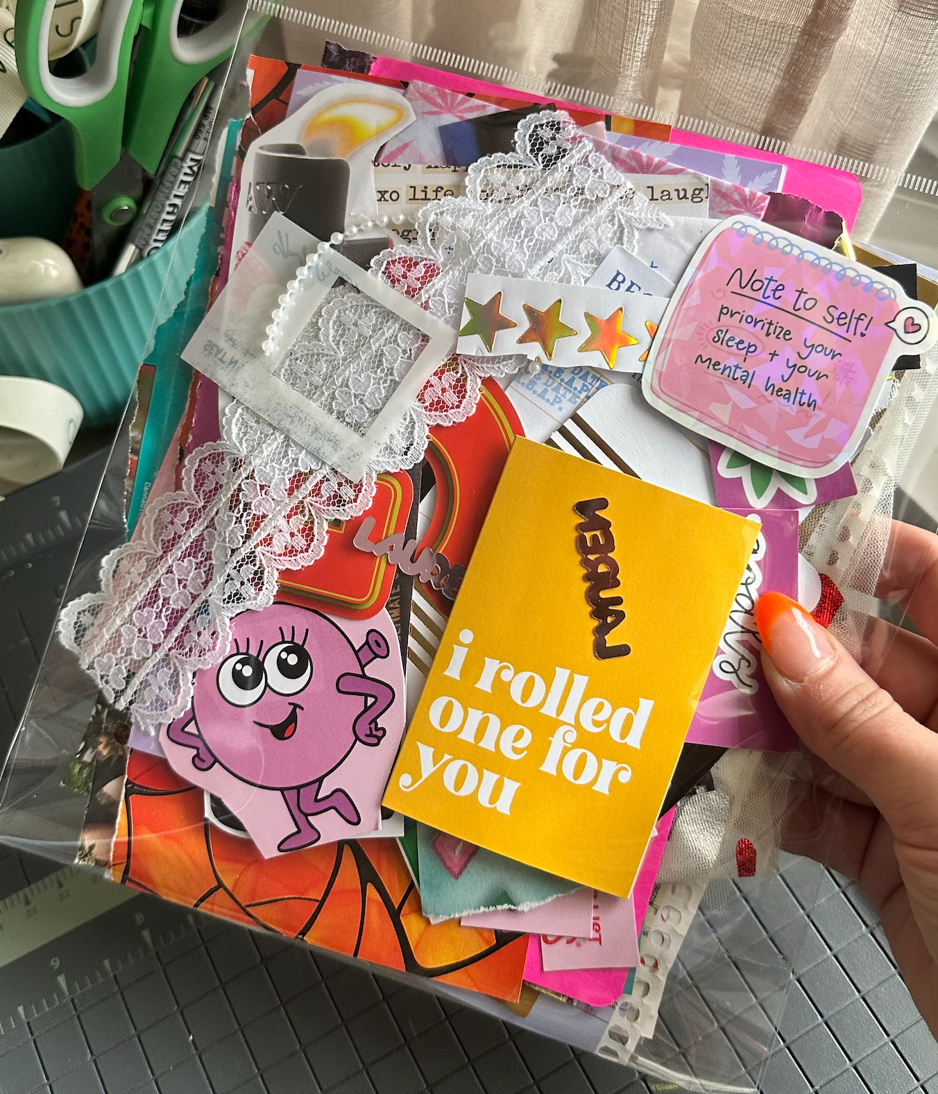 Collection of colorful paper cutouts and a hand holding a yellow card with text, on a table with crafting tools.