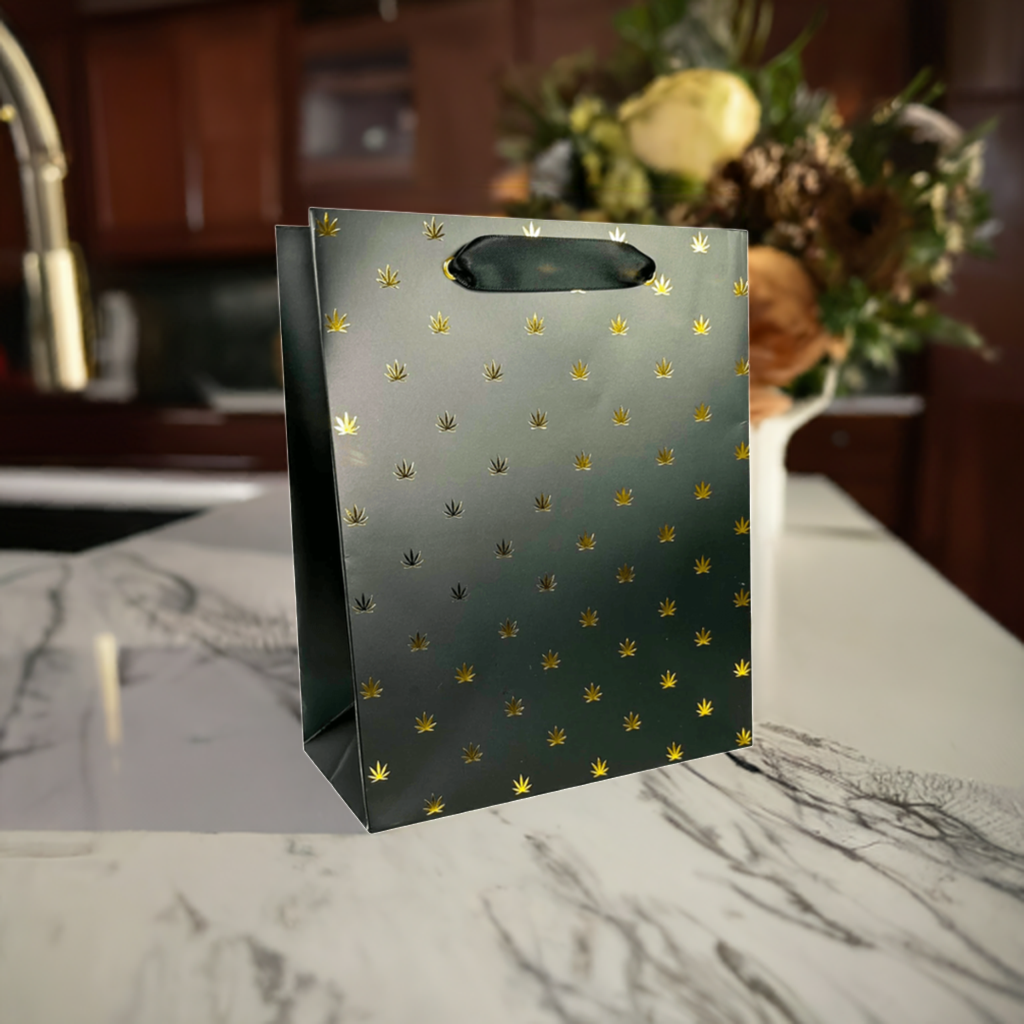 Black gift bag with gold foil cannabis leaf pattern and black ribbon handles.