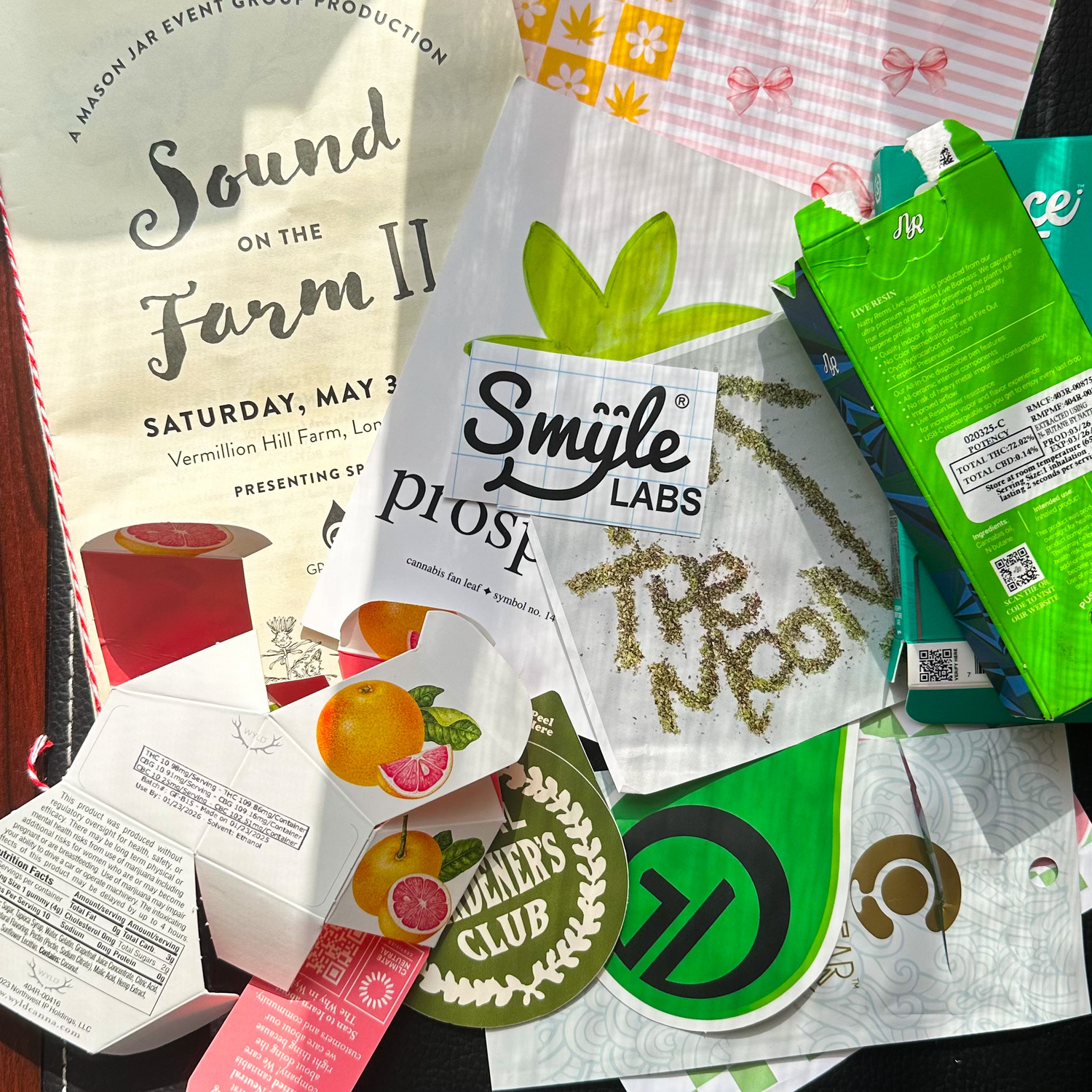 A colorful flat lay of junk journal scraps, including cannabis edible packaging, concert flyers, fruit illustrations, branded exit bags, and paper cutouts—showcasing a creative collection of upcycled materials for junk journaling.