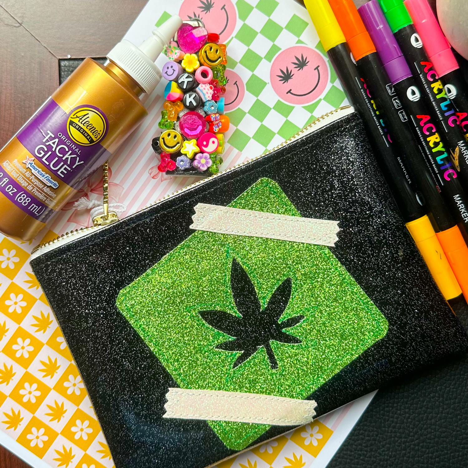 Flat lay of a glittery cannabis-themed stash bag surrounded by markers, tacky glue, smiley beads, and KushKards prints—perfect for organizing junk journal supplies.