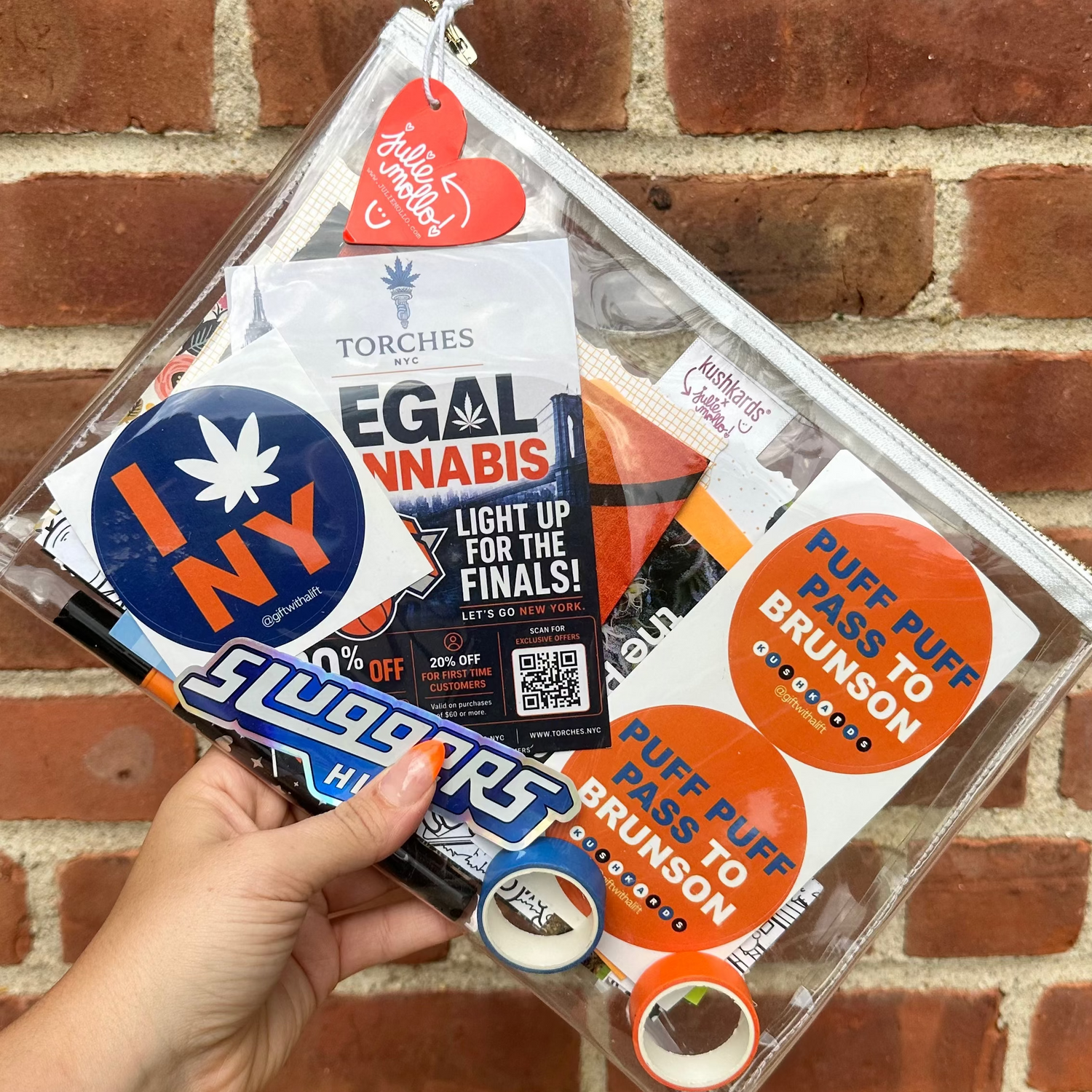 Hand holding a clear clutch bag with various stickers against a brick wall.