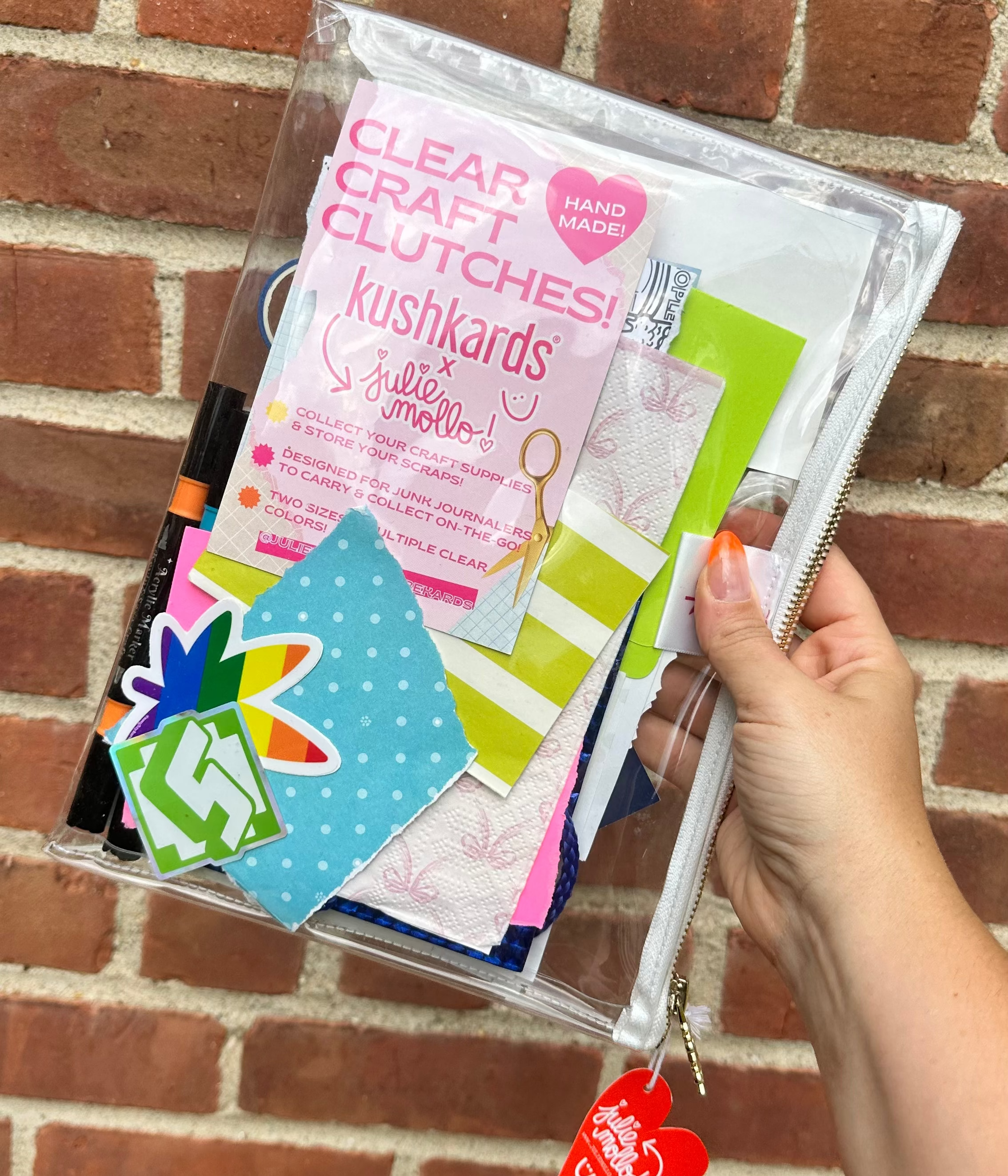 Clear craft clutch with colorful items against a brick wall
