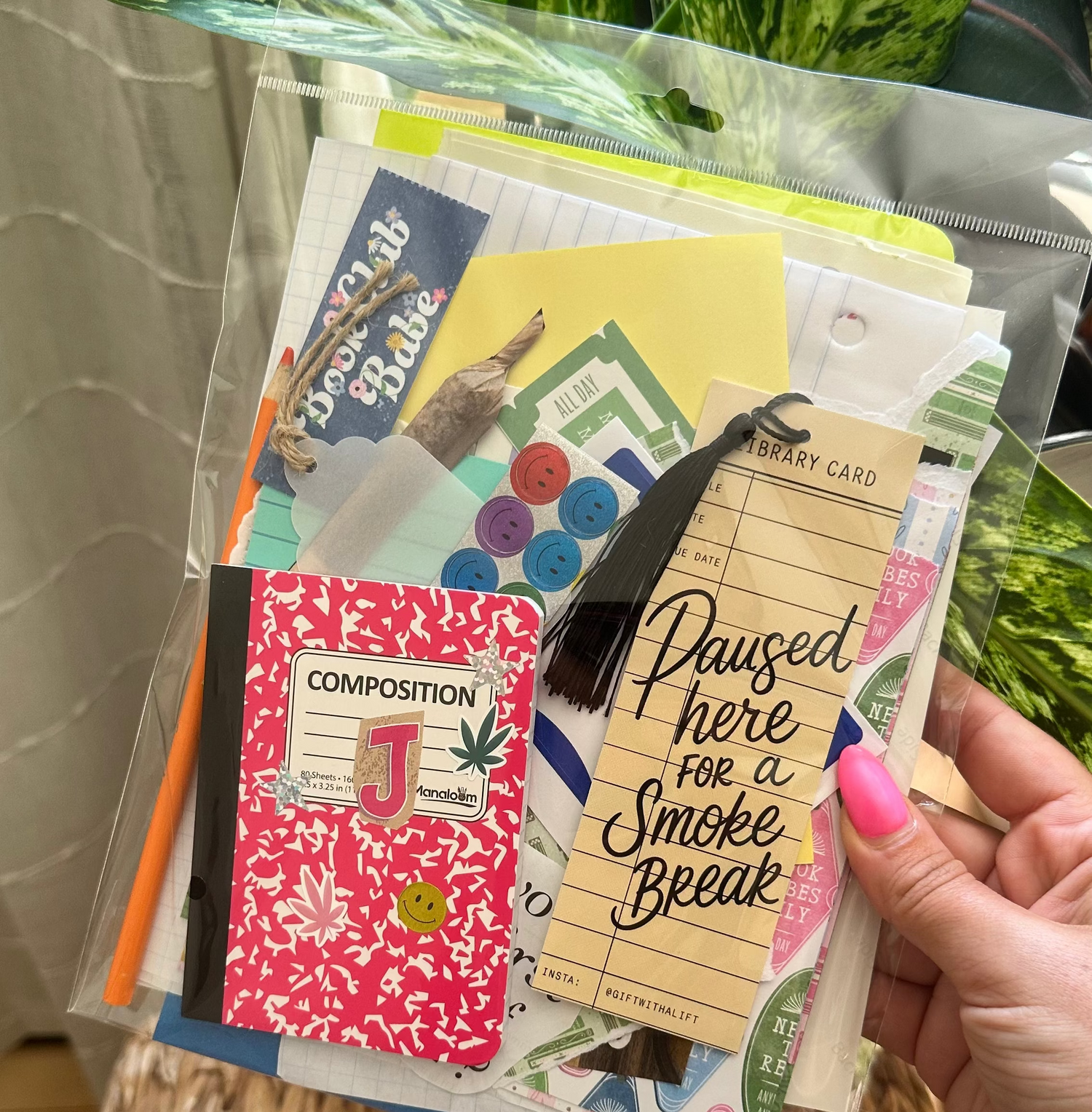 A hand with pink nail polish holds a clear plastic bag filled with assorted stationery items, including a pink floral composition notebook, a yellow card reading &