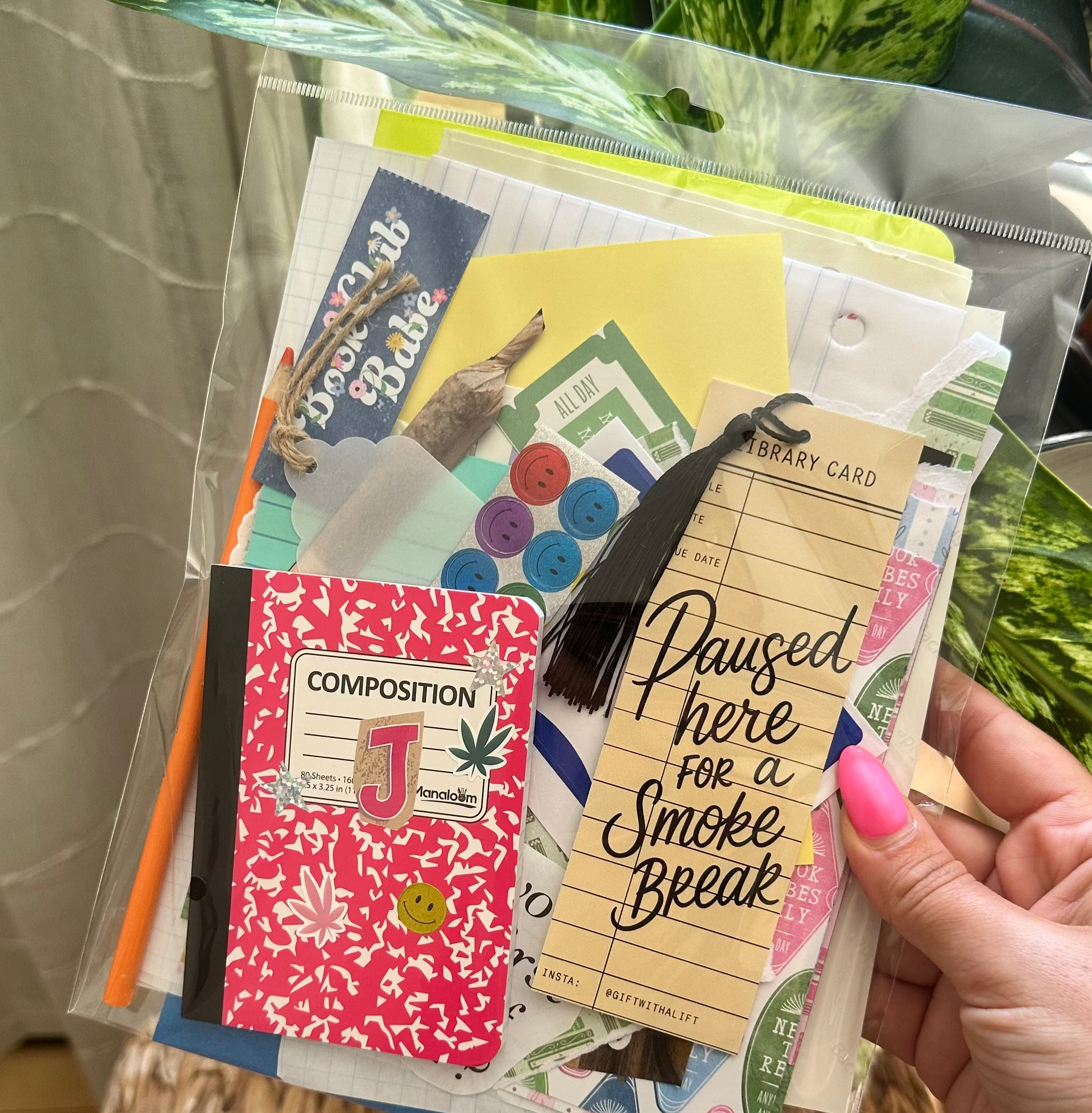 A hand with pink nail polish holds a clear plastic bag filled with assorted stationery items, including a pink floral composition notebook, a yellow card reading &