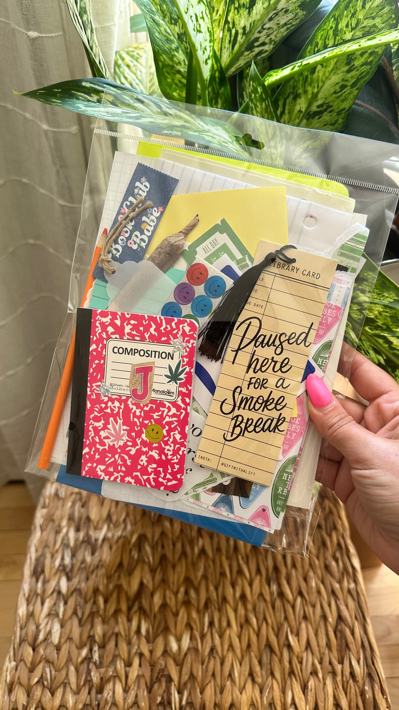 A hand with pink nail polish holds a clear plastic bag filled with assorted stationery items, including a pink floral composition notebook, a yellow card reading 'Paused here for a Smoke Break,' and other colorful paper goods, set against a background of green plants and a woven basket.