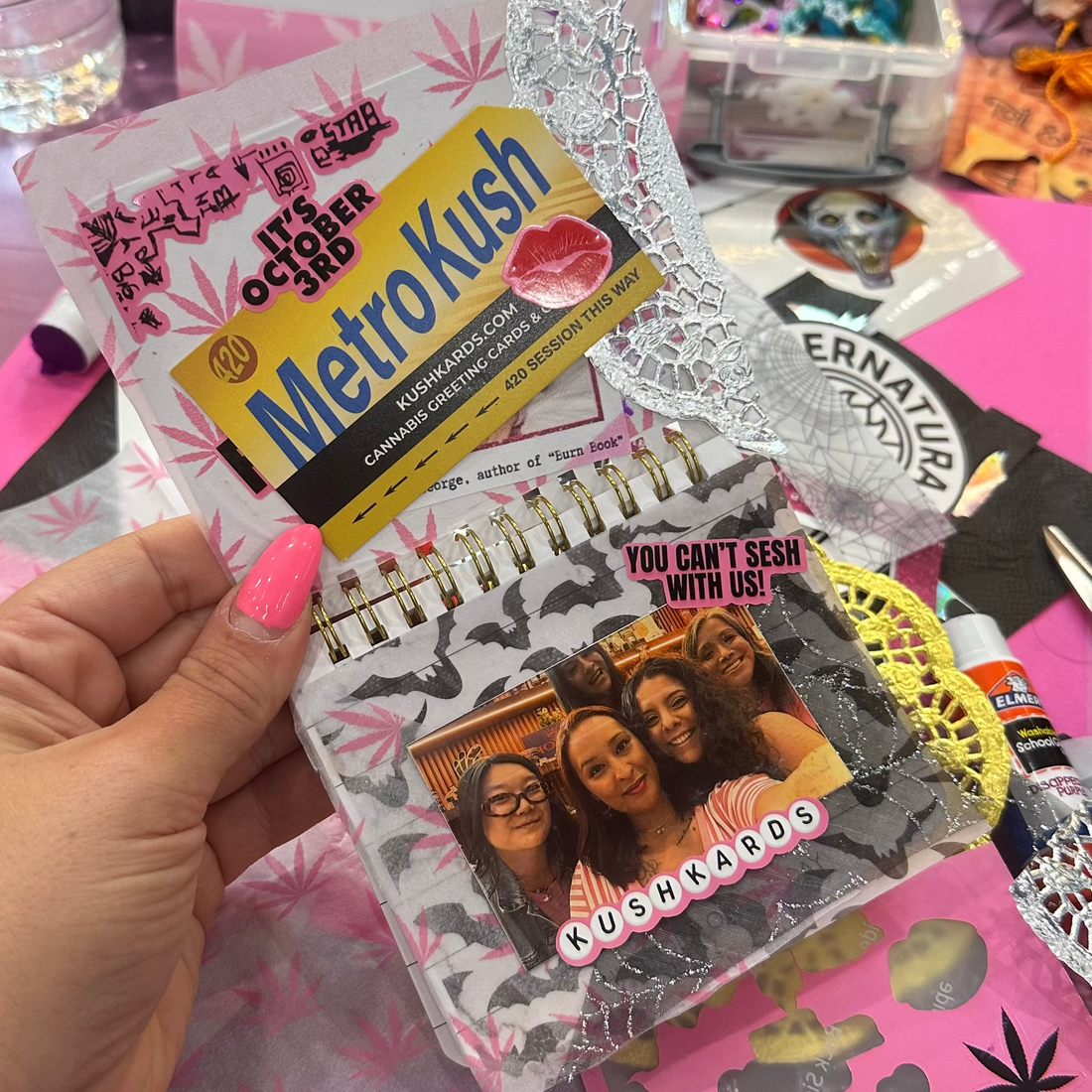 A hand with pink nail polish holds a small zippered notebook or journal. The cover features a collage of photos, a yellow and black &