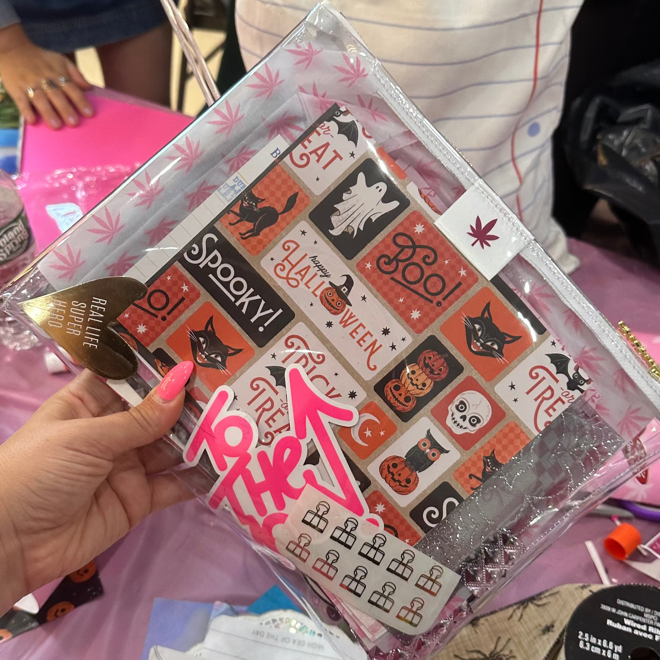 A hand with pink nail polish holds a clear zip pouch filled with Halloween-themed stickers, including &