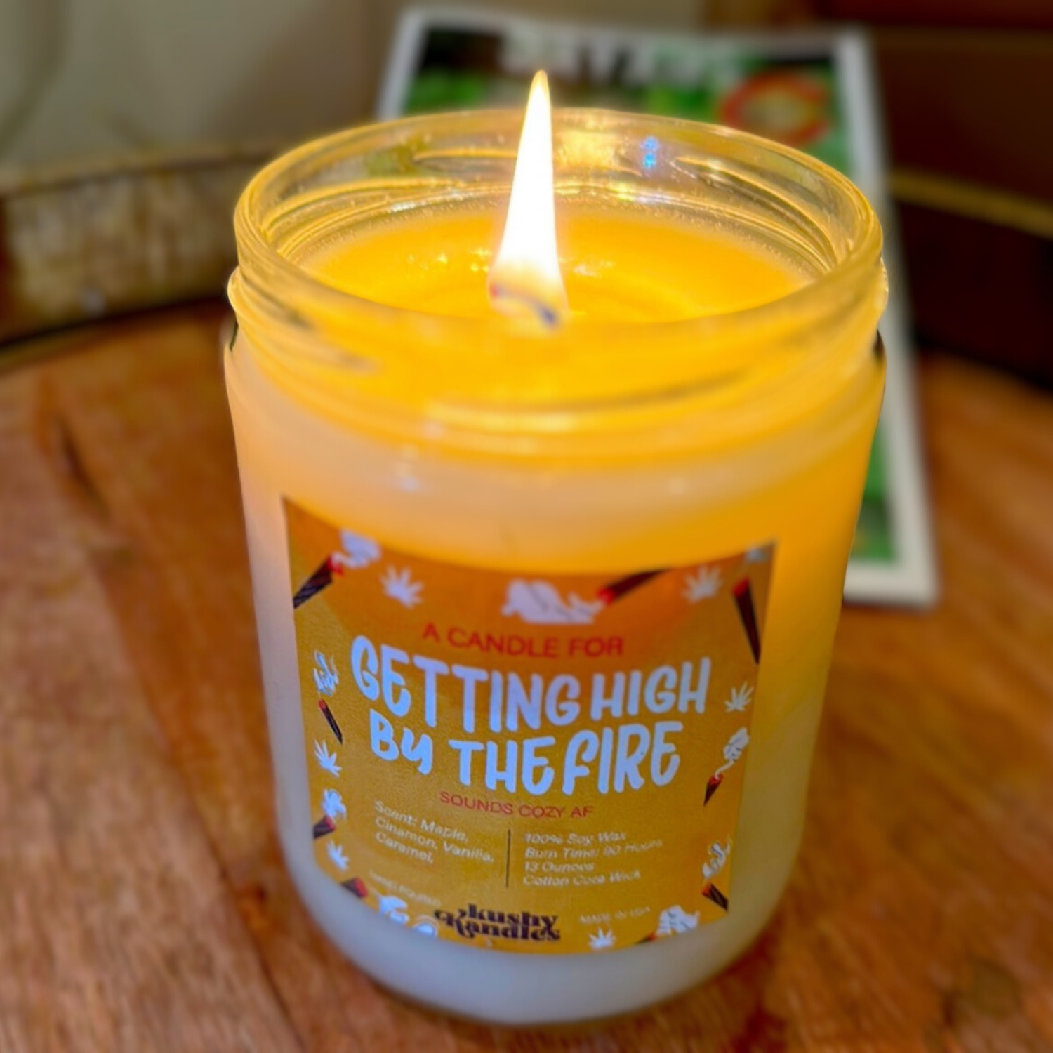 A glowing candle labeled &quot;Getting High by the Fire&quot; on a wooden surface. The candle is lit, showing its warm maple, cinnamon, vanilla, and caramel scent, creating a cozy and relaxing atmosphere.