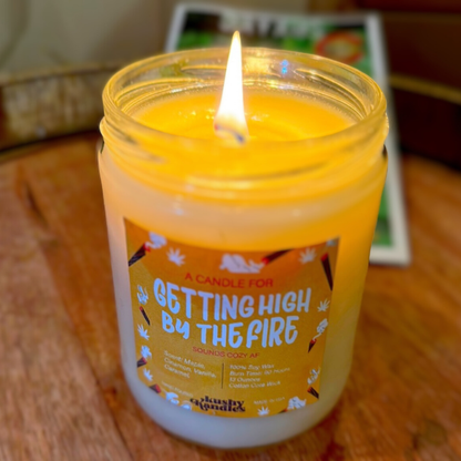 A glowing candle labeled &quot;Getting High by the Fire&quot; on a wooden surface. The candle is lit, showing its warm maple, cinnamon, vanilla, and caramel scent, creating a cozy and relaxing atmosphere.