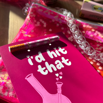 A close-up of a greeting card with a bright pink front featuring the playful phrase "I'd hit that" above a stylized illustration of a bong. A matching one-hitter pipe is attached. In the foreground, there's a decorative pen with pink and white patterns and glittery brown accents, lying atop the card. Behind the card is a Kush Clutch, a translucent pink bag filled with pot leaf-shaped confetti and adorned with a chunky clear chain. The items are presented on a wooden surface in a sunlit setting.