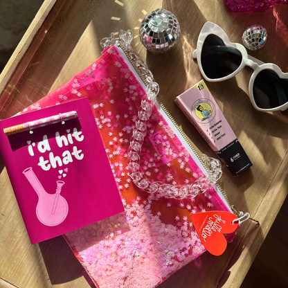 An assorted collection of items arranged on a wooden surface, highlighted by natural sunlight. The collection includes a bright pink greeting card with the phrase 'i'd hit that' and an illustration of a bong, a Kush Clutch with a clear pink surface filled with pot leaf confetti and a chunky clear  chain, a small disco ball, stylish white sunglasses with black lenses. The scene conveys a sense of fun and contemporary lifestyle.