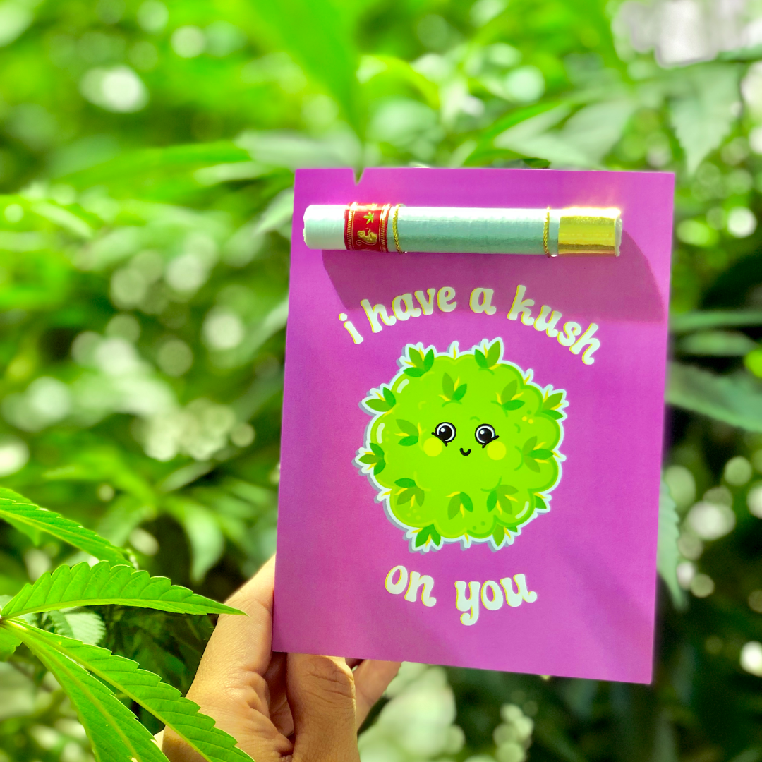 A hand holding a vibrant purple greeting card featuring a cute, smiling cannabis leaf and the pun &quot;I have a Kush on You.&quot; The card is set against a lush, green, leafy background.