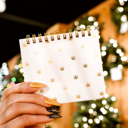 Hand holding a Pot Leaf Print Pocket Spiral Notebook with gold spiral and printed pot leaves against a festive background.