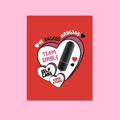 A humorous greeting card with a red background featuring playful black and white candy hearts with phrases like 'WE SUCKED ANYWAY,' 'TEAM SINGLE,' 'SELF CARE,' and 'CUPID STUPID.' A black vibrator is prominently attached, contributing to the card's theme of self-love and independence.