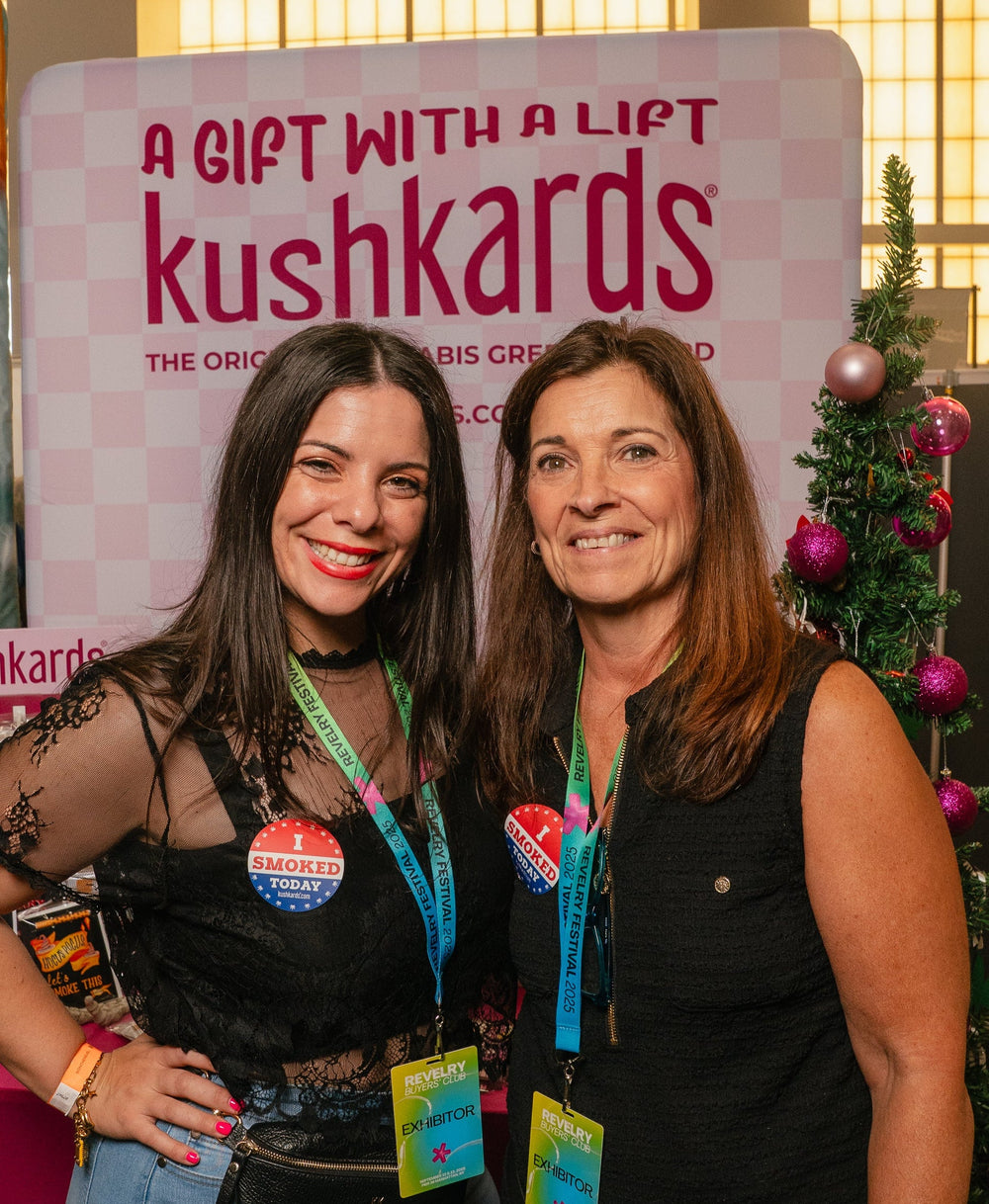 Two women smiling in front of a Kushkards event booth at an indoor convention. One woman wears a black lace top and light blue jeans, and the other wears a black sleeveless top.