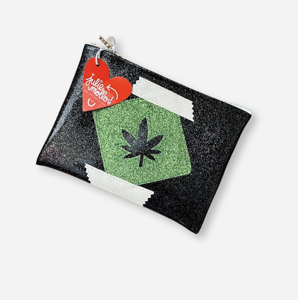 A black glittery pouch with a green pot leaf cut out of the center. The pouch has a zipper closure and a red heart-shaped tag that reads &quot;Julie Mollo&quot; attached to the zipper pull.